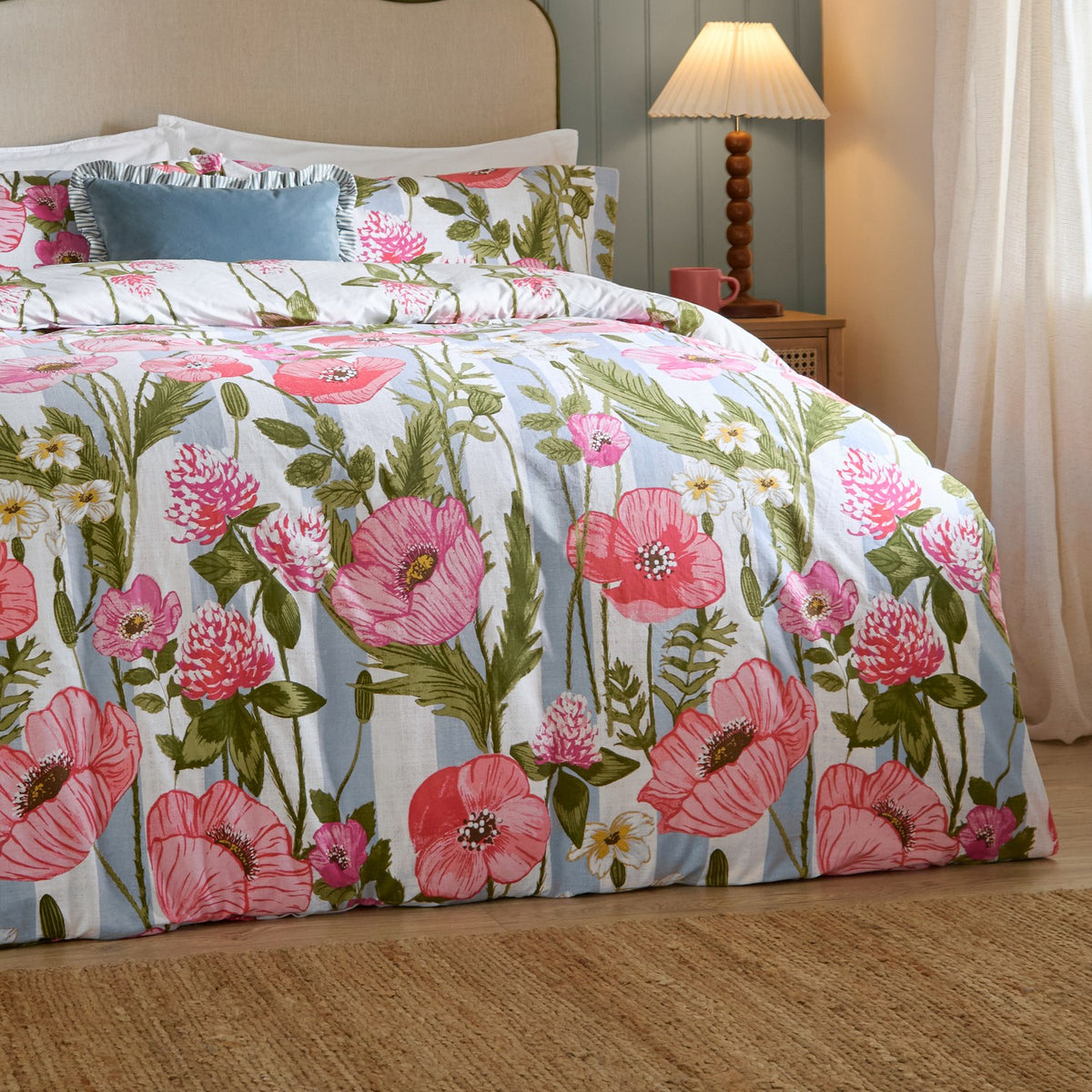 Poppy Trail Reversible Floral Duvet Cover Set in Multi on a bed, showing pink and coral poppies with green foliage on a striped ground, styled in a bedroom with a blue lamp on a bedside table.
