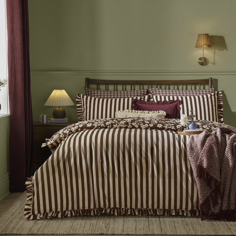 Posie Reversible Floral Duvet Cover Set in chocolate showing the striped side with frilled trim, styled on a bed with floral cushions in a green bedroom.