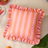 Palm Royale Stripe Ruffle square cushion in pink and orange, featuring bold vertical stripes and a matching ruffled edge trim, shown outdoors on a woven mat.