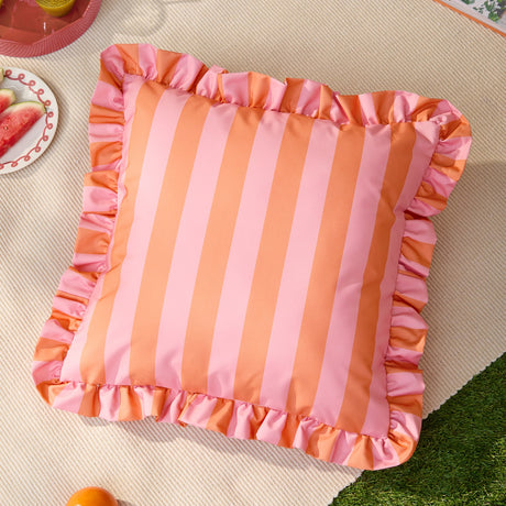 Palm Royale Stripe Ruffle square cushion in pink and orange, featuring bold vertical stripes and a matching ruffled edge trim, shown outdoors on a woven mat.