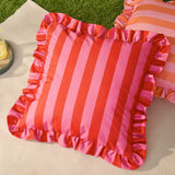 Palm Royale Stripe Ruffle Printed Polyester Cushion in pink and red vertical stripes with a gathered ruffle trim, shown on a light rug outdoors near a striped pillow.