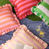 Palm Royale Stripe Ruffle Printed Polyester Cushion in pink and red stripes with gathered ruffle trim, shown among matching striped cushions in an outdoor garden setting.
