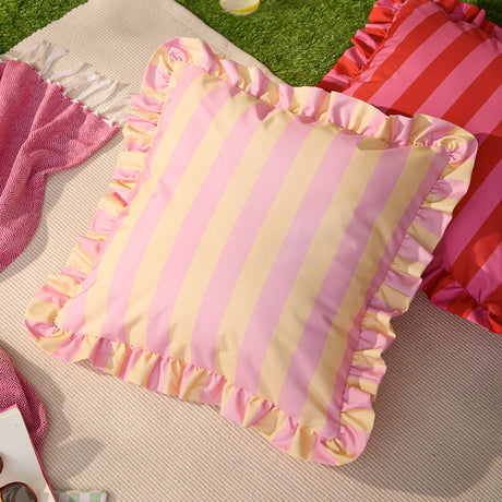 Palm Royale Stripe Ruffle square cushion in pink and yellow vertical stripes with a matching gathered ruffle trim, shown outdoors on a picnic blanket with a red striped cushion nearby.