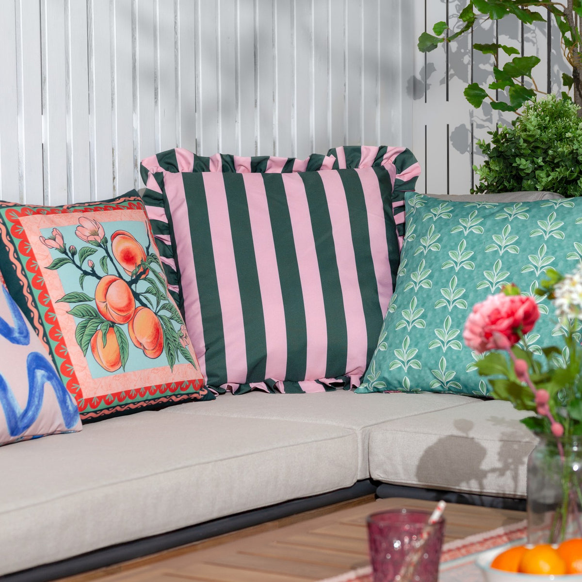 Palm Royale Stripe polyester cushion in forest green and pink with ruffle trim, styled on an outdoor sofa alongside patterned scatter cushions.