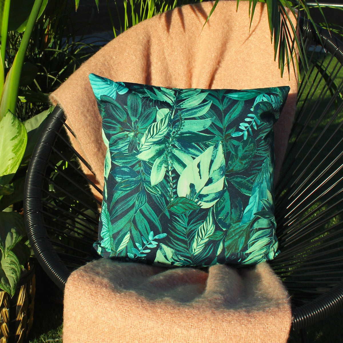 Square outdoor cushion, 43 x 43cm, with a dense tropical leaf print in deep green and blue-green tones on a near-black ground, shown on a chair with foliage behind.