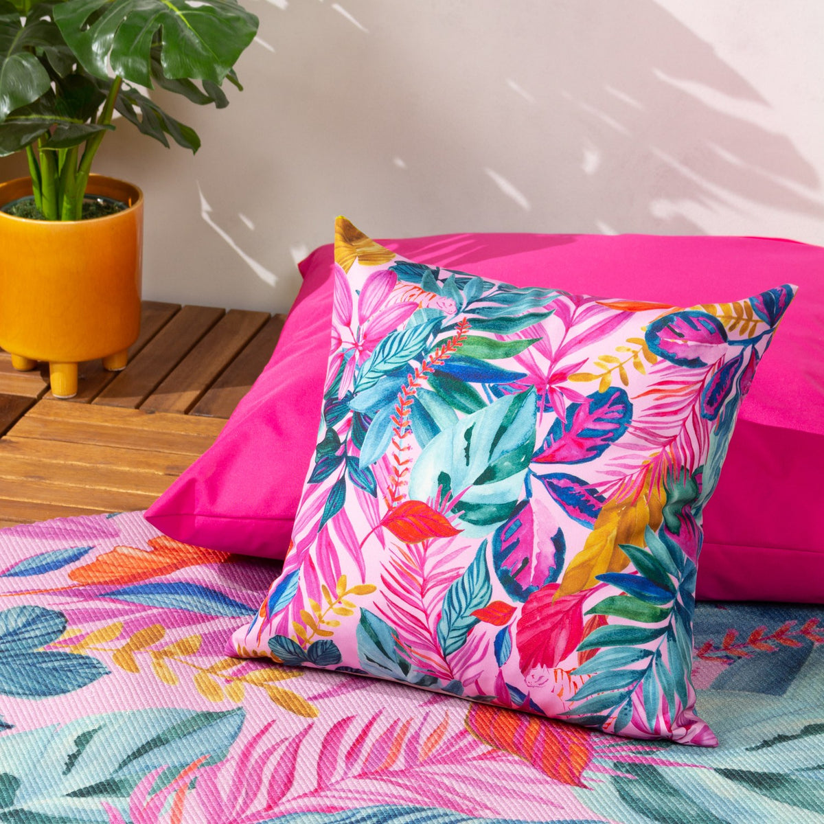 Psychedelic Jungle Reversible Polyester Cushion in multi, 43 x 43cm square, with a tropical leaf print in pink, teal, green and gold, shown on a pink cushion in an outdoor patio setting.