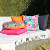 Psychedelic Jungle reversible polyester cushion in a pink, teal and orange tropical print, styled with pink, orange and dark grey cushions on an outdoor sofa.