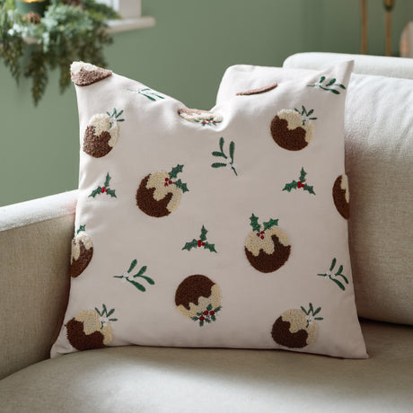 Square cream velvet cushion with tufted Christmas pudding motifs in brown and cream, accented by embroidered holly, mistletoe, and red berries, shown on a sofa.