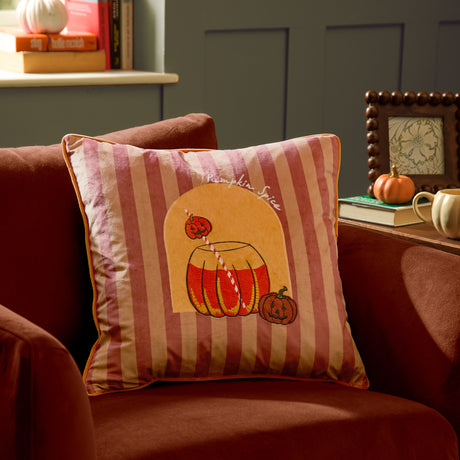 Pumpkin Spice Striped Velvet Cushion in orange on a red sofa, showing an embroidered pumpkin drink and jack-o'-lanterns on berry and terracotta stripes with orange piping.