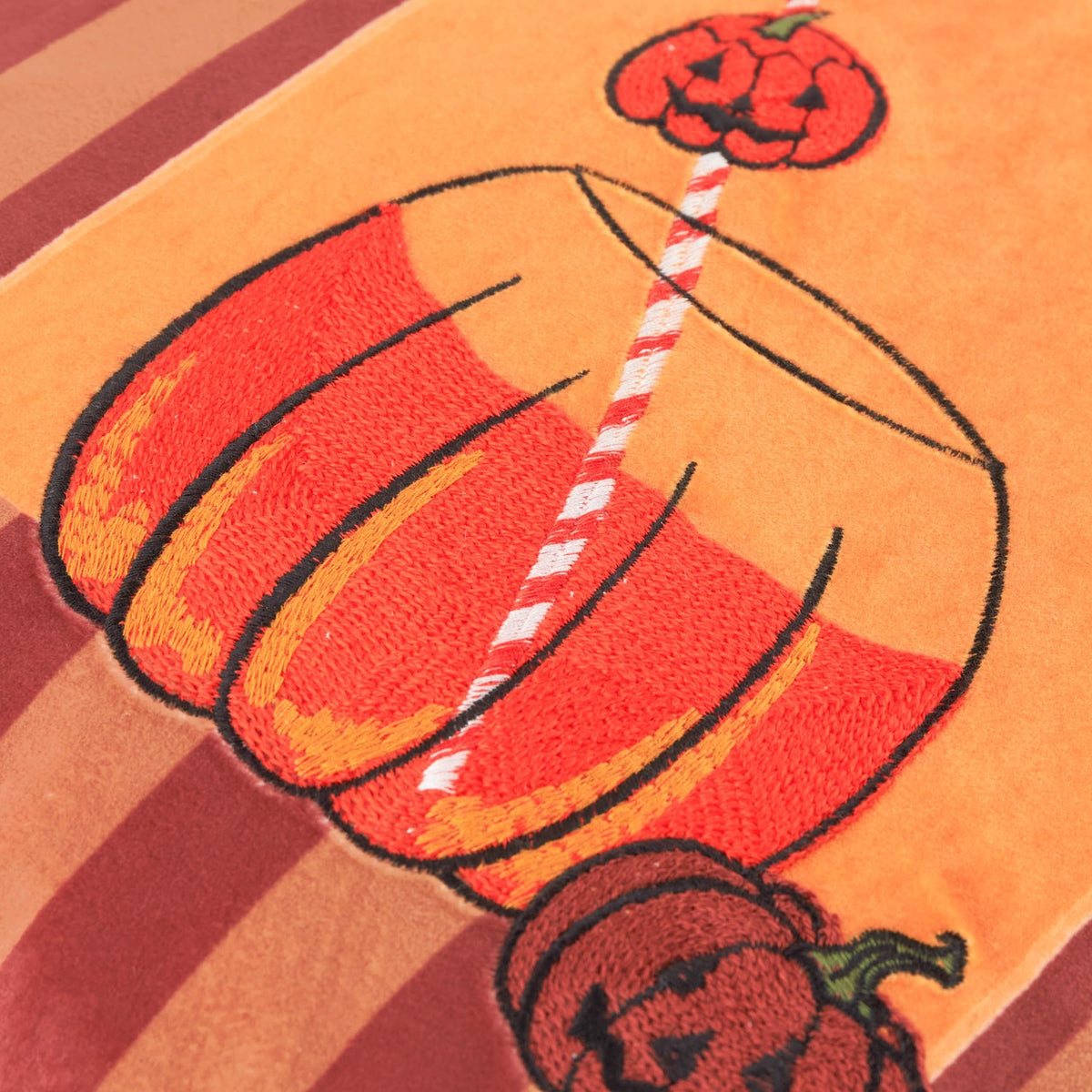 Close-up of the Pumpkin Spice Striped Velvet Cushion in orange, showing embroidered jack-o'-lantern pumpkins and a striped straw in a drink glass on a berry and terracotta striped ground.
