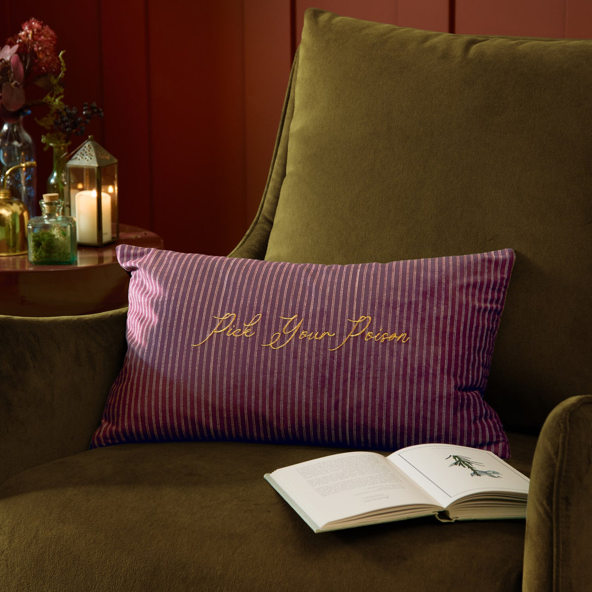 Rectangular purple striped velvet cushion with "Pick Your Poison" in gold embroidery, styled on an olive green armchair beside an open book.