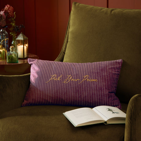 Rectangular purple striped velvet cushion with "Pick Your Poison" in gold embroidery, styled on an olive green armchair beside an open book.