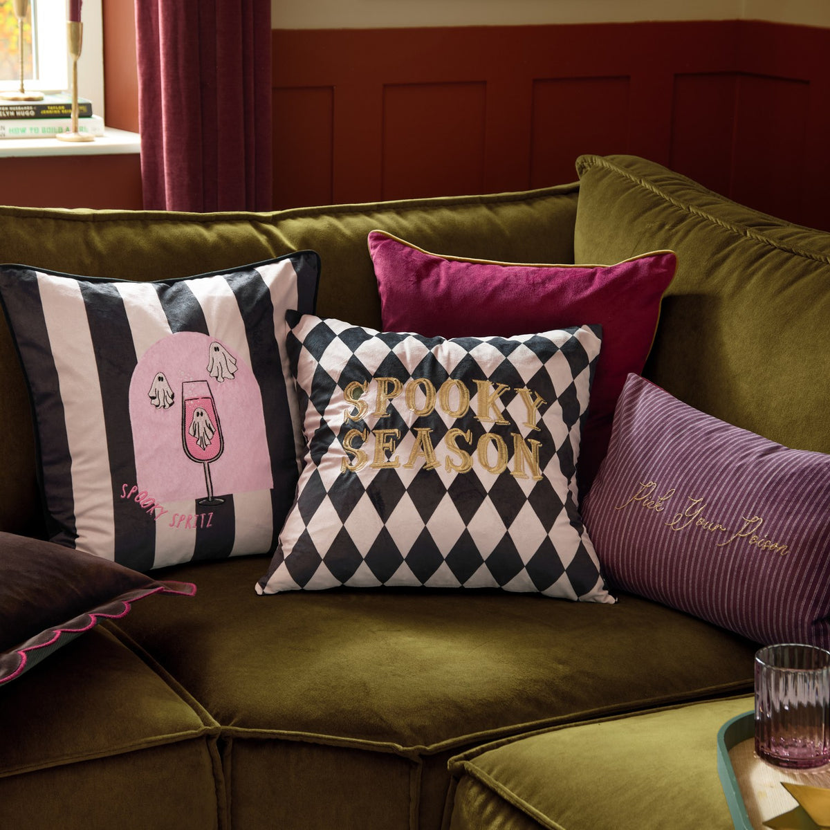 Rectangular striped velvet cushion in plum and mauve with "Pick Your Poison" gold embroidered text, styled on an olive sofa among patterned Halloween cushions.