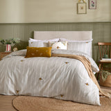 Rambbes Animal Velvet Cushion in honey gold with embroidered bee and leaf motifs, styled on a bed with striped bee-print bedding in a green-panelled bedroom.