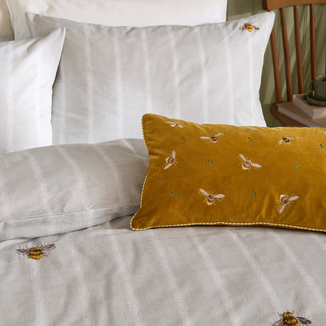 Ramble Cotton Striped Duvet Cover Set in Natural, with grey striped bedding and embroidered bee motifs, styled with a mustard velvet bee-print bolster cushion on a bed.