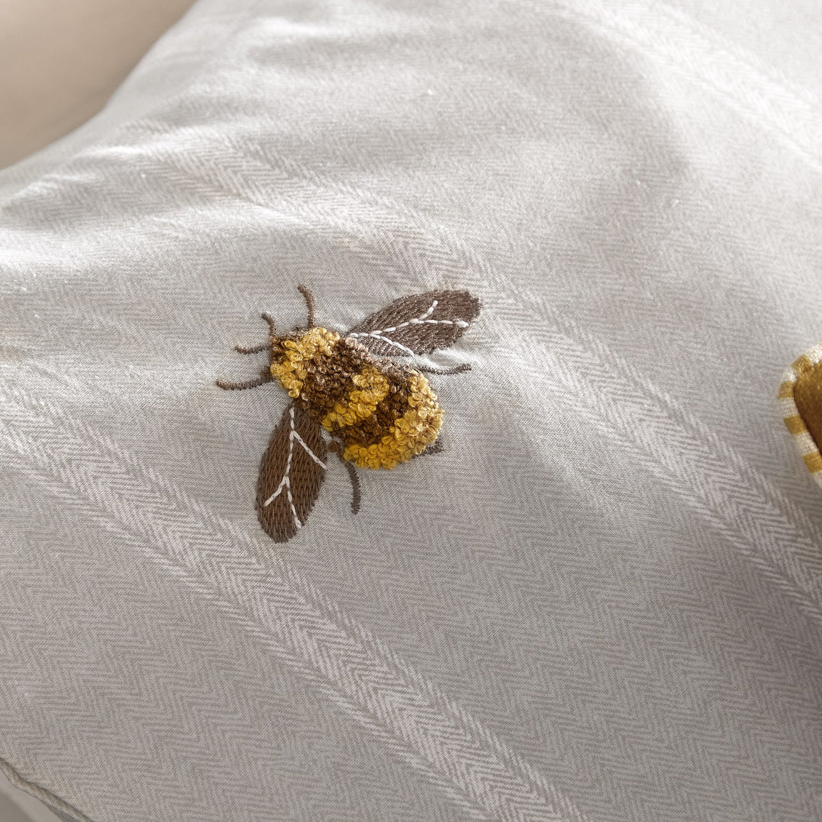 Close-up of the Ramble Cotton Striped Duvet Cover Set in natural, showing an embroidered, tufted bee in yellow, gold and brown thread on a subtle striped cotton ground.