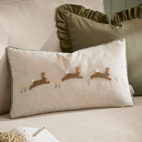 Rectangular Rambhar cushion in natural linen with tufted leaping hares, pom tails, embroidered flowers, and gingham piping, shown on a sofa with sage green ruffled cushions behind.
