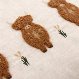 Close-up of the Rambhco Velvet Linen Cushion in Ginger, showing tufted rust-brown highland cows in curly looped yarn with embroidered grass tufts on an oatmeal-flecked linen ground.
