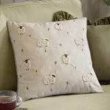 Rambshe Velvet Linen Cushion in Natural, a square cushion with tufted sheep and embroidered grass on a flecked linen-blend face, shown on a sofa with green cushions.