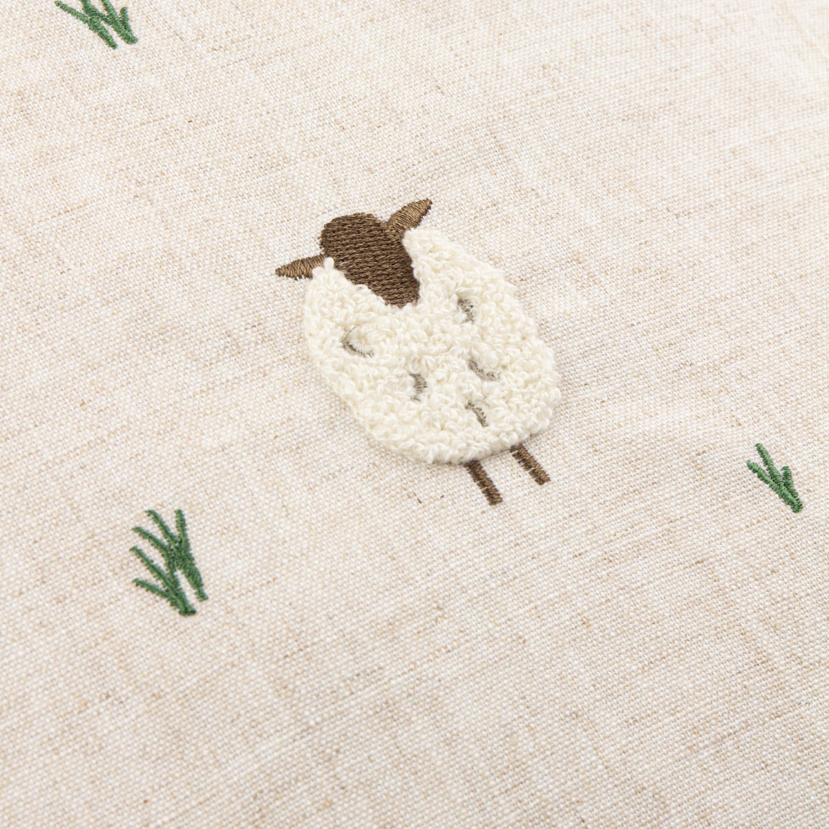 Close-up of the Rambshe Velvet Linen Cushion in natural, featuring a tufted sheep with looped white body, embroidered brown head and legs, and green grass stitching on flecked linen-blend fabric.