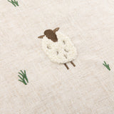 Close-up of the Rambshe Velvet Linen Cushion in natural, featuring a tufted sheep with looped white body, embroidered brown head and legs, and green grass stitching on flecked linen-blend fabric.
