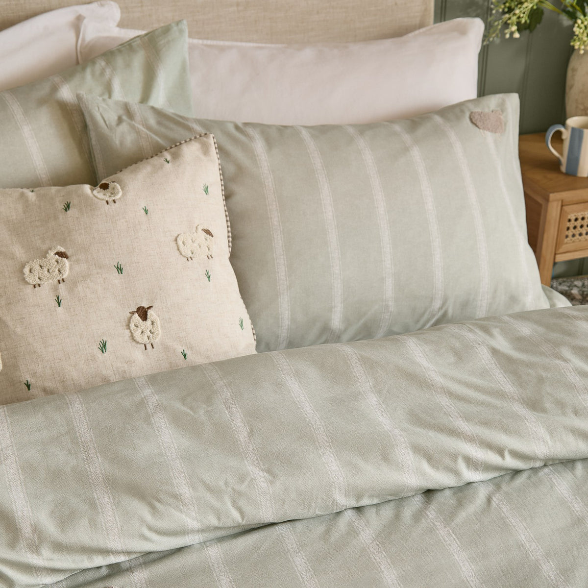 Rambshe Velvet Linen Cushion in Natural with tufted sheep and grass design on a flecked ground, shown on a bed styled with striped green bedding and pillows.