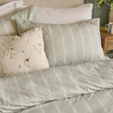 Rambshe Velvet Linen Cushion in Natural with tufted sheep and grass design on a flecked ground, shown on a bed styled with striped green bedding and pillows.