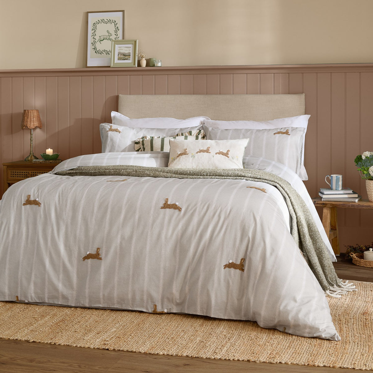 Ramble Cotton Animal Duvet Cover Set in Sage on a bed, featuring embroidered hare motifs on a striped white and beige ground, styled with white pillowcases and a green throw.
