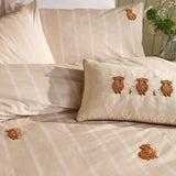 Ramble cotton duvet cover set in natural beige with cream stripes, featuring tufted rust-brown highland cow embroidery, styled with a matching bolster cushion on a bed.