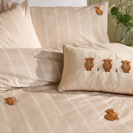 Ramble cotton duvet cover set in natural beige with cream stripes, featuring tufted rust-brown highland cow embroidery, styled with a matching bolster cushion on a bed.