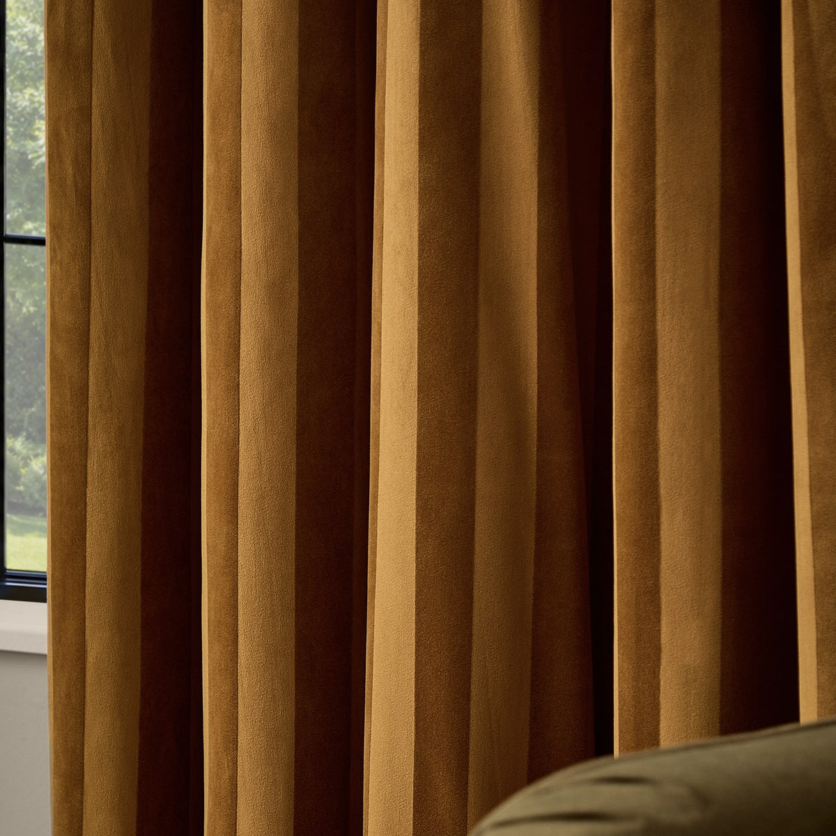 Regency Stripe Velvet Fully Lined Eyelet Curtains in antique gold, shown in deep, light-catching folds beside a window, with an olive sofa edge visible in the room.
