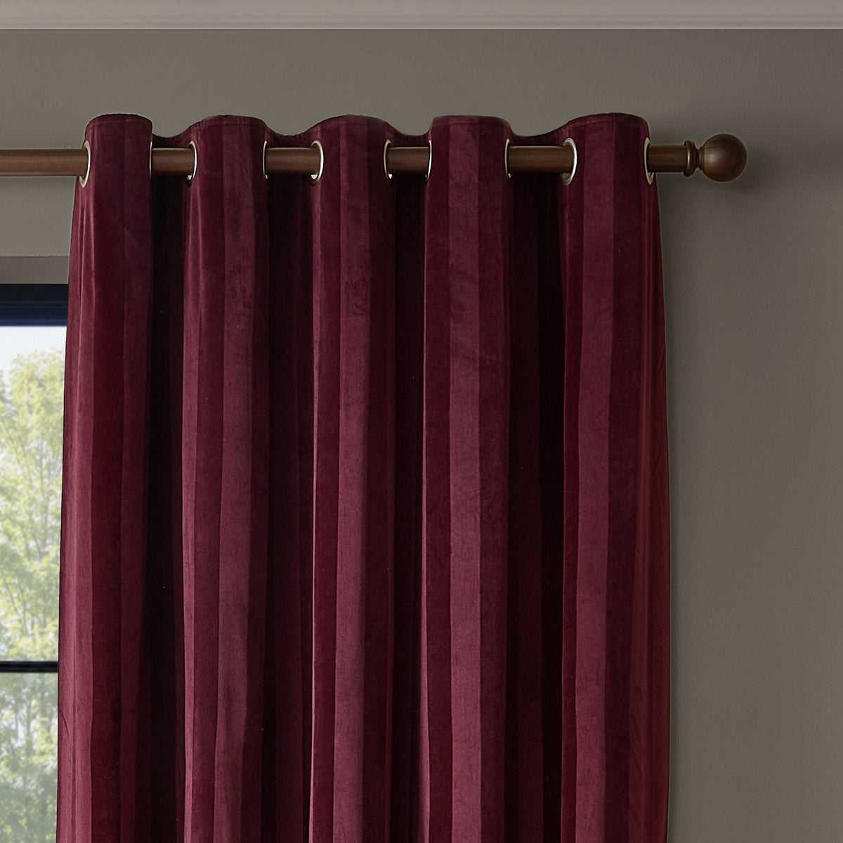 Regency Stripe Velvet Fully Lined Eyelet Curtains in burgundy, self-striped velvet fabric with grommet top hanging from a wooden pole against a beige wall.