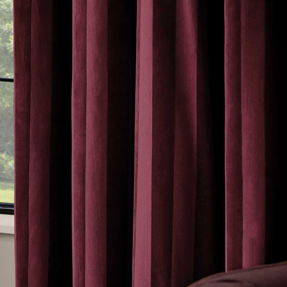Close-up of Regency Stripe velvet eyelet curtains in burgundy, showing deep folds with a subtle self-stripe catching light across the pile, beside a window.