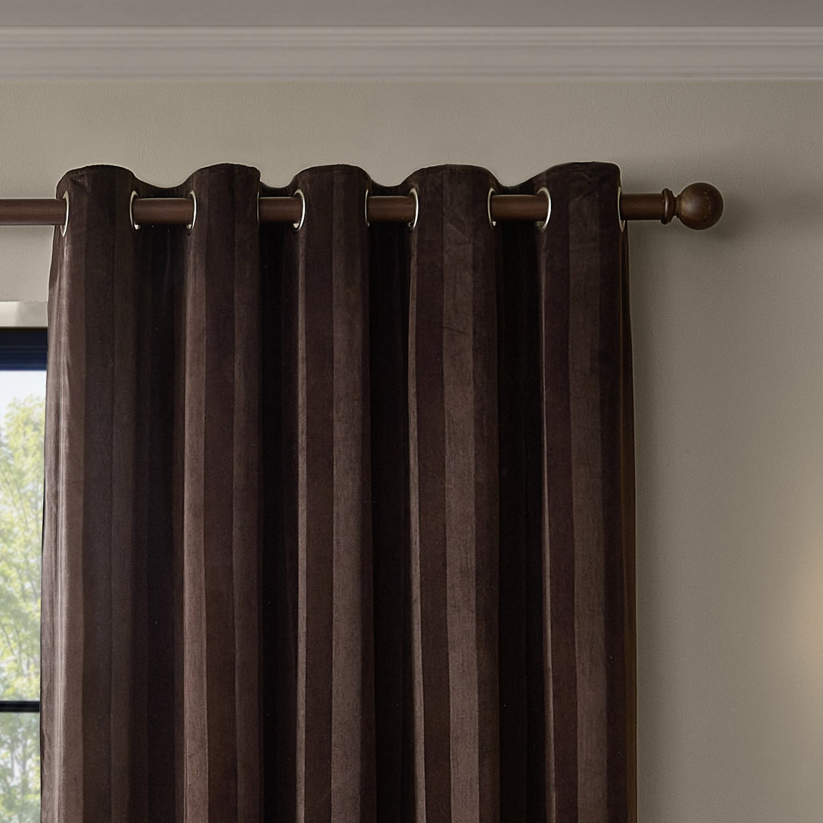 Regency Stripe Velvet Fully Lined Eyelet Curtains in chocolate brown, showing vertical pile stripes and metal eyelet heading on a wooden pole, hung by a window.