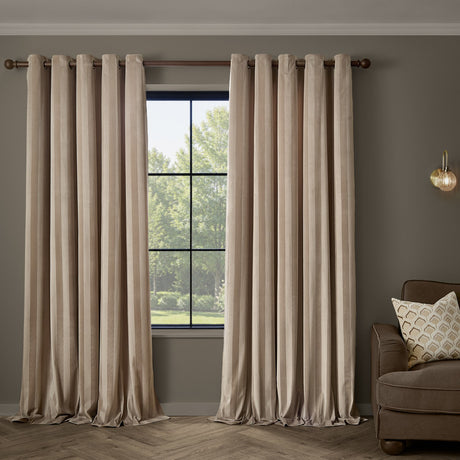 Regency Stripe Velvet Fully Lined Eyelet Curtains in beige, self-striped pile, hung from a wooden pole in a living room beside a window and brown armchair.