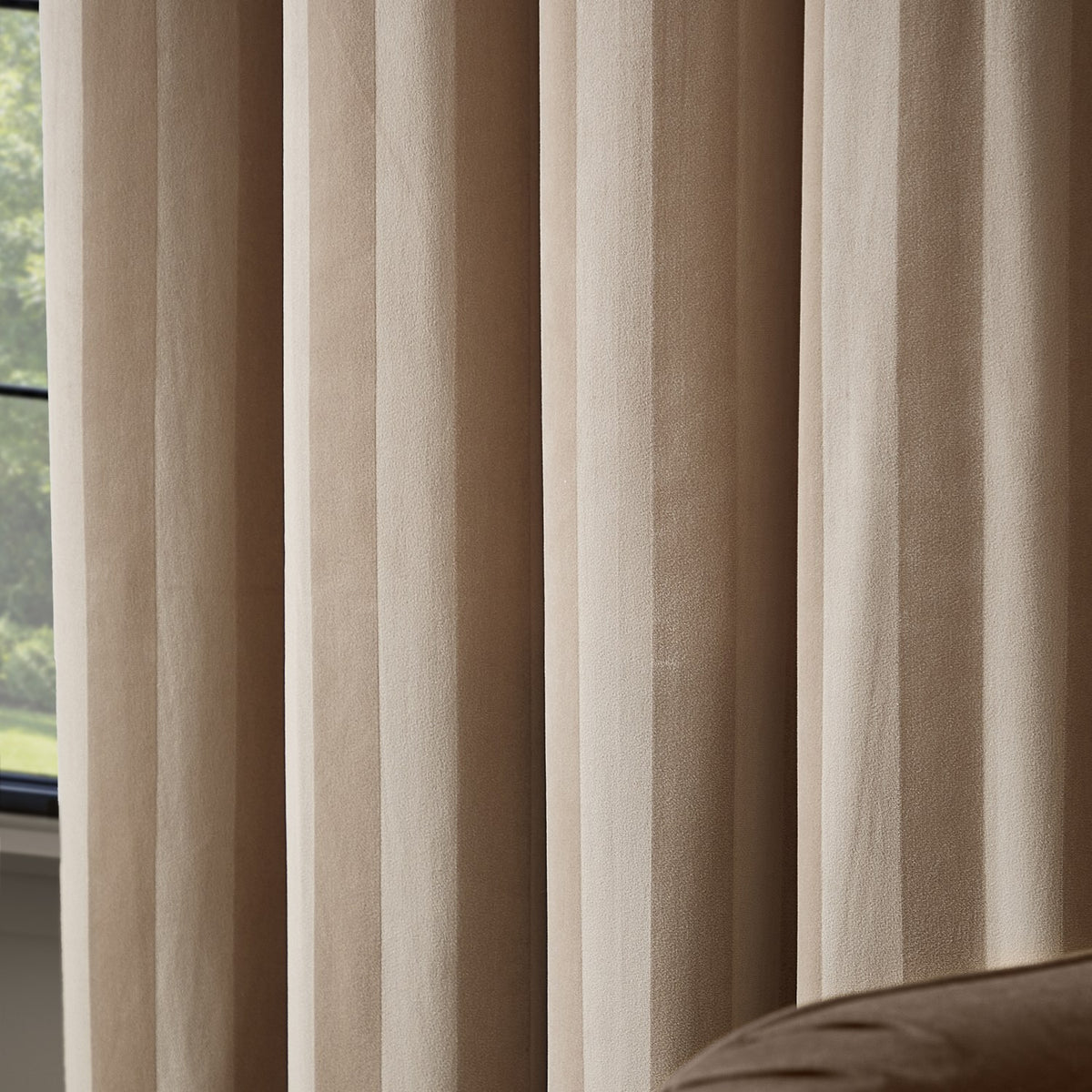 Regency Stripe fully lined eyelet curtains in beige velvet with a subtle self-stripe pile, shown floor-to-ceiling beside a window in a living room setting.
