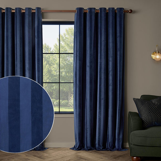 Regency Stripe Velvet Fully Lined Eyelet Curtains in Midnight navy blue, shown on a copper rod in a living room with a tonal stripe swatch inset and a green armchair nearby.