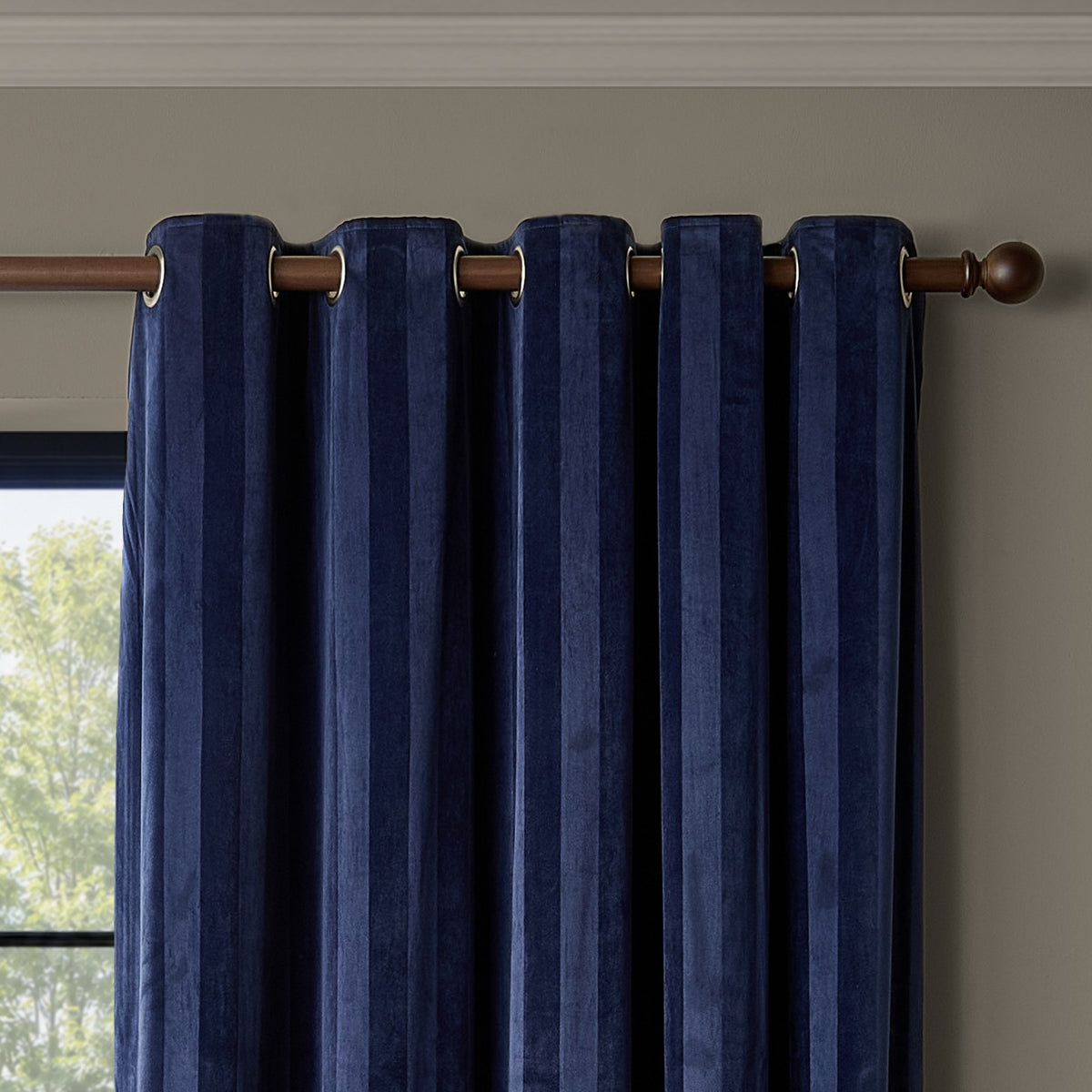 Midnight navy velvet eyelet curtains with subtle vertical stripe and fully lined panels, hung on a dark wood pole with round finials against a neutral wall.