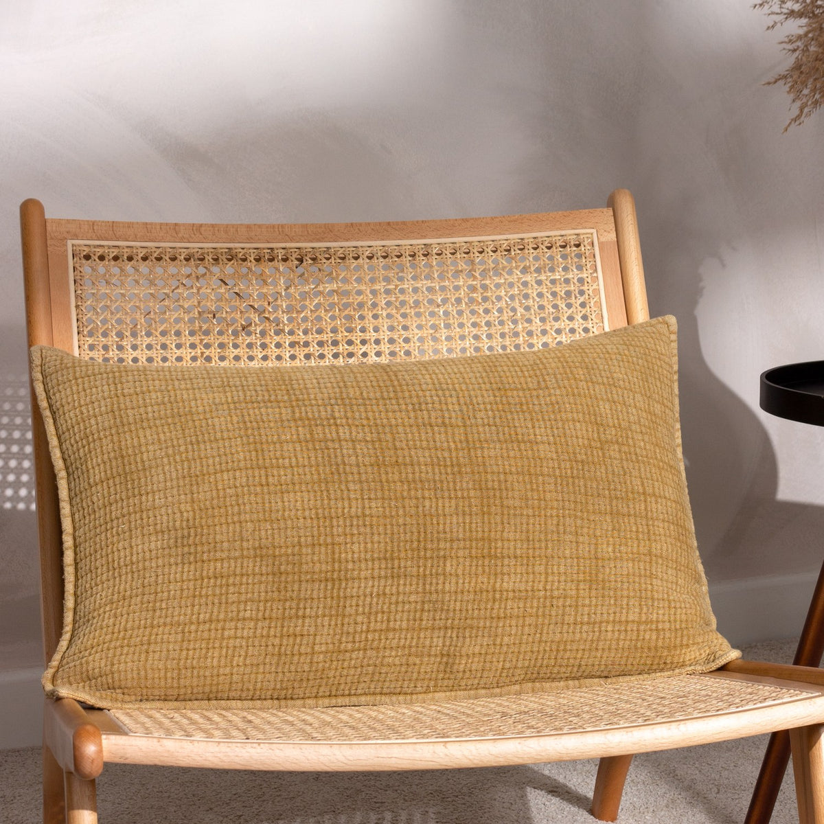 Ribble Reversible Cotton Cushion in honey, a rectangular bolster-style cushion with a ribbed woven texture, shown propped on a rattan cane chair.