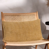 Ribble Reversible Cotton Cushion in honey, a rectangular bolster-style cushion with a ribbed woven texture, shown propped on a rattan cane chair.