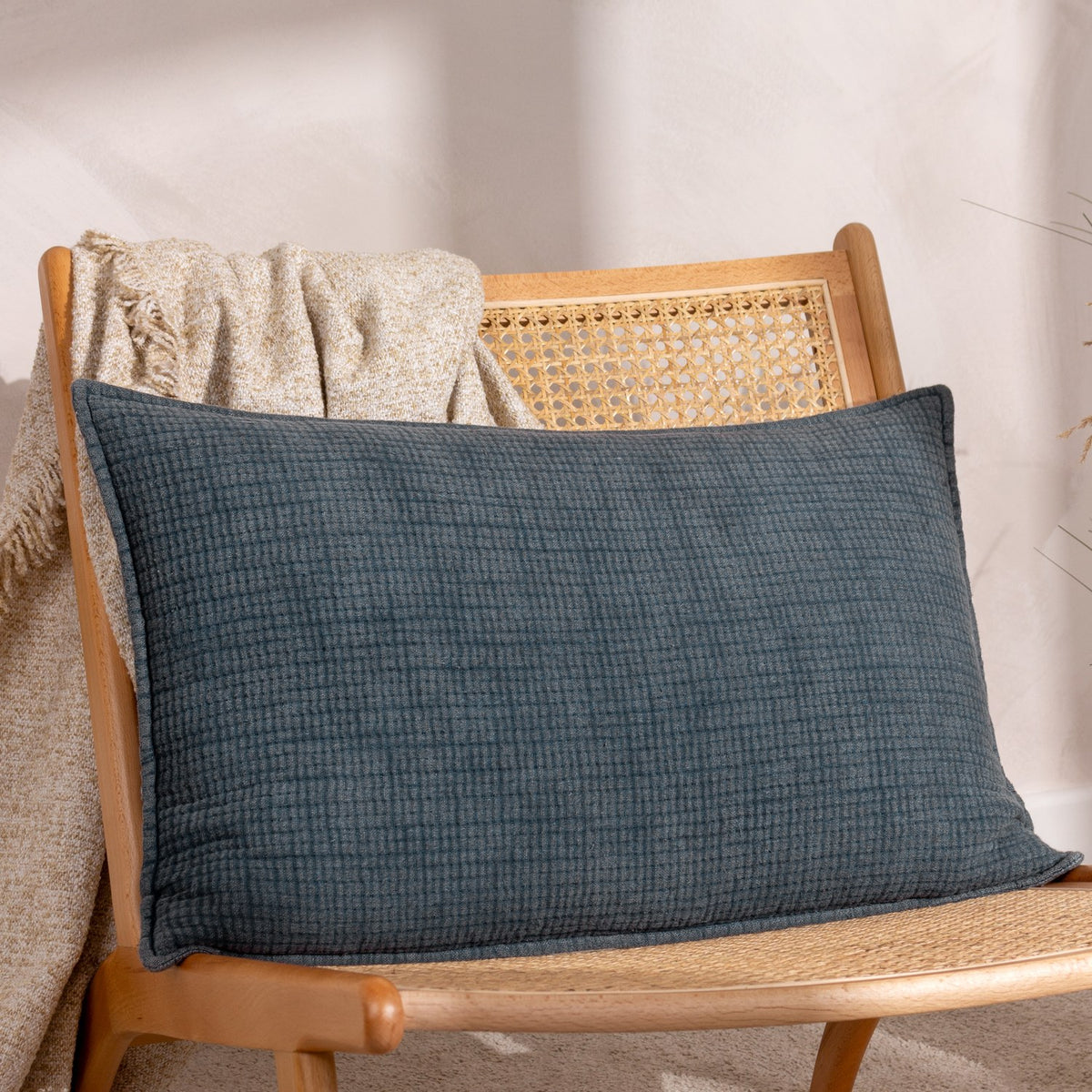 Ribble Reversible Cotton Cushion in ink blue, rectangular bolster shape with ribbed woven texture, styled on a cane-back wooden chair with a beige throw.