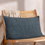 Ribble Reversible Cotton Cushion in ink blue, rectangular bolster shape with ribbed woven texture, styled on a cane-back wooden chair with a beige throw.