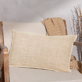 Ribble Reversible Cotton Cushion in natural with a textured ribbed weave, shown on a wooden armchair beside a woven throw and dried branches.