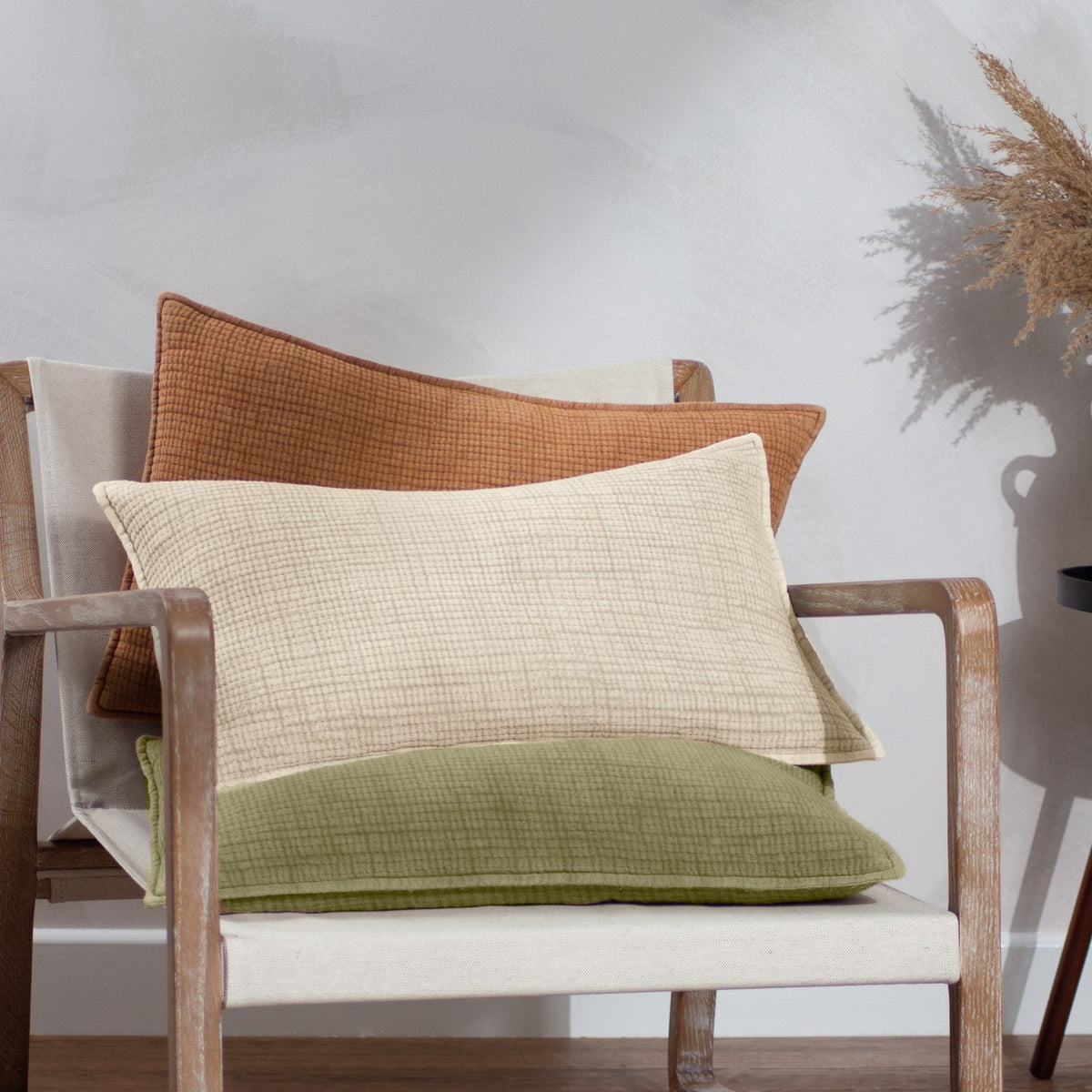 Ribble Reversible Cotton Cushion in natural cream shown with rust and olive green cushions, textured ribbed weave, on a wooden armchair with linen upholstery.
