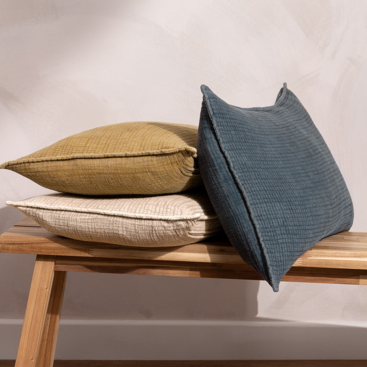 Ribble Reversible Cotton Cushion in natural, shown with mustard and slate blue textured cushions stacked on a wooden bench against a light background.