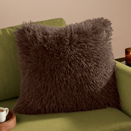 Rowan Velvet Faux Fur Cushion in chocolate brown, shown with its shaggy Mongolian-style pile face on a green sofa in a living room setting.