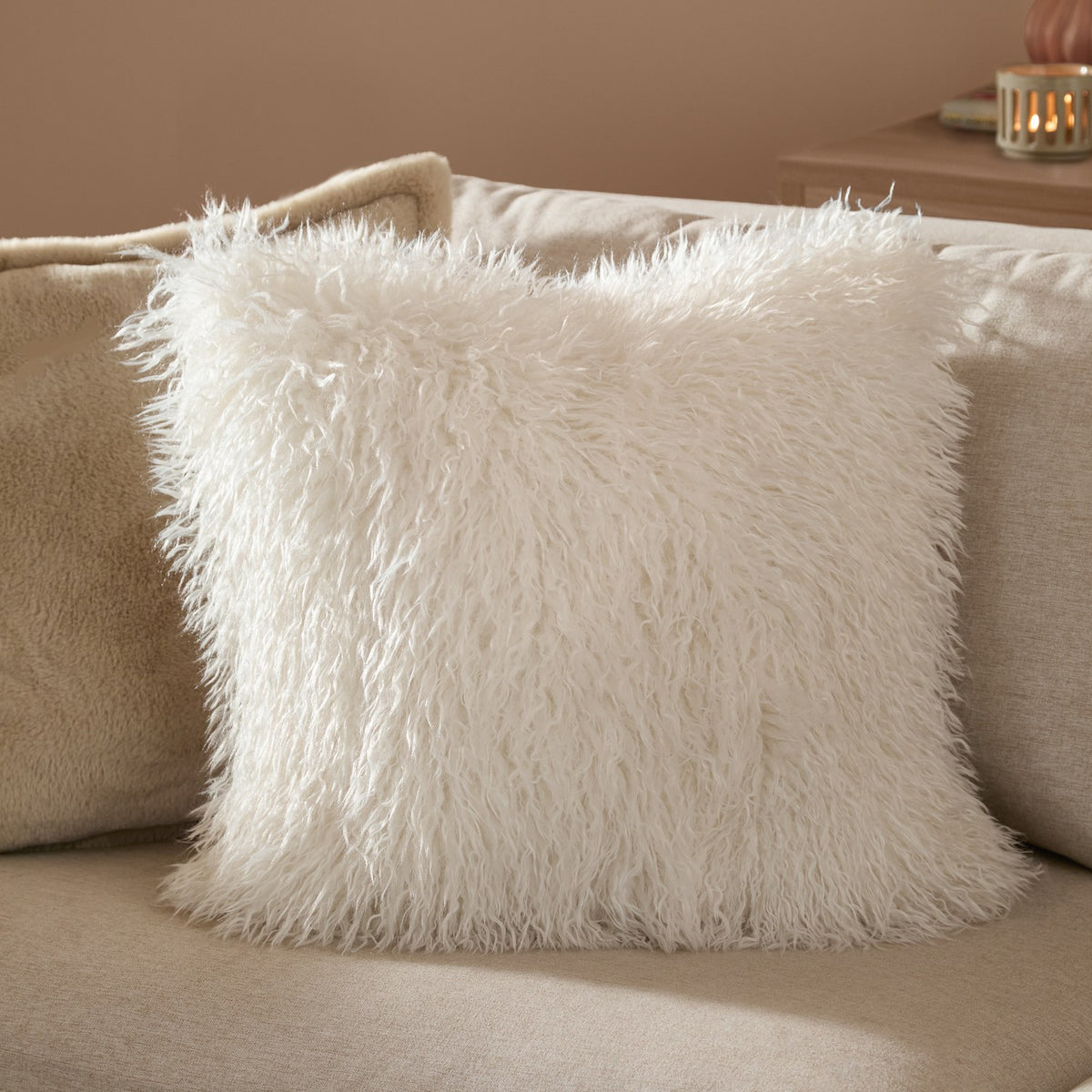 Rowan Faux Fur Plain Velvet Cushion in ecru, square scatter cushion with shaggy long-pile front, shown on a beige sofa in a living room setting.