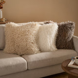 Rowan Faux Fur Cushion in ecru with shaggy Mongolian-style pile, shown among similar textured cushions on a beige sofa in a living room setting.