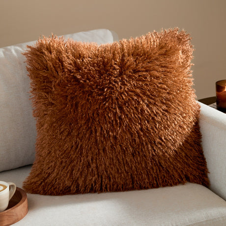 Rowan Faux Fur Plain Velvet Cushion in Ginger with shaggy, long-pile fur front, shown square-shaped on a light grey sofa in a living room setting.