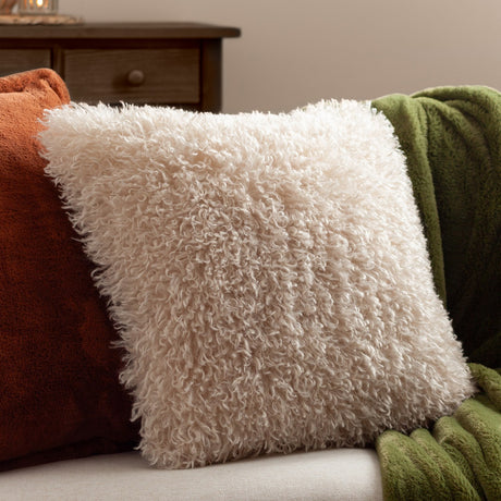 Rowan Faux Fur Polyester Velvet Cushion in natural cream with curly shaggy pile, square 50x50cm, shown on a sofa beside orange and green throws.